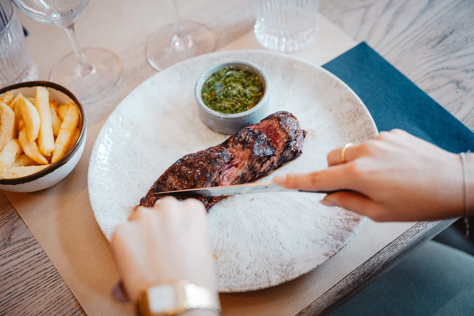 Een gast snijdt een steak aan, met frietjes en chimichurri ernaast