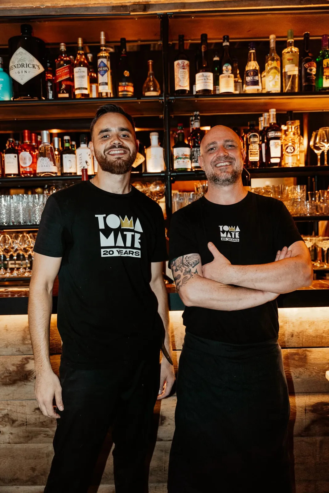 Twee medewerkers in Toma Maté-shirts poseren voor de bar met flessen
