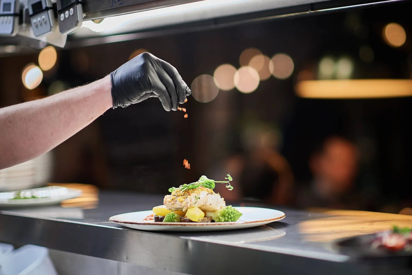 Een kok met zwarte handschoen werkt een bord af aan de pass