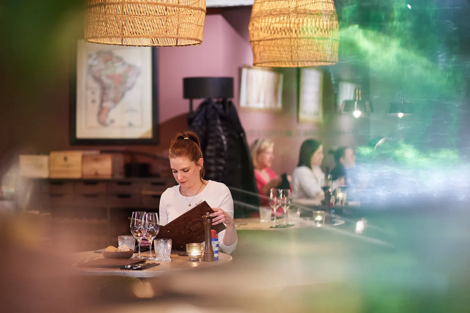 Een gaste leest de menukaart aan tafel onder rotan lampen