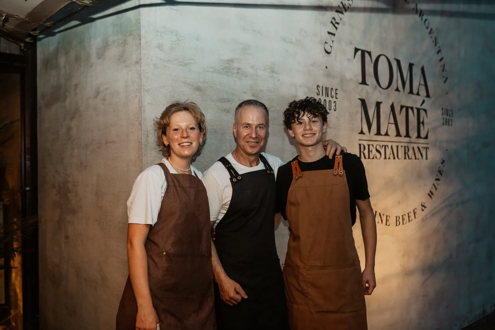 Drie teamleden in schort voor de muur met het Toma Maté-logo