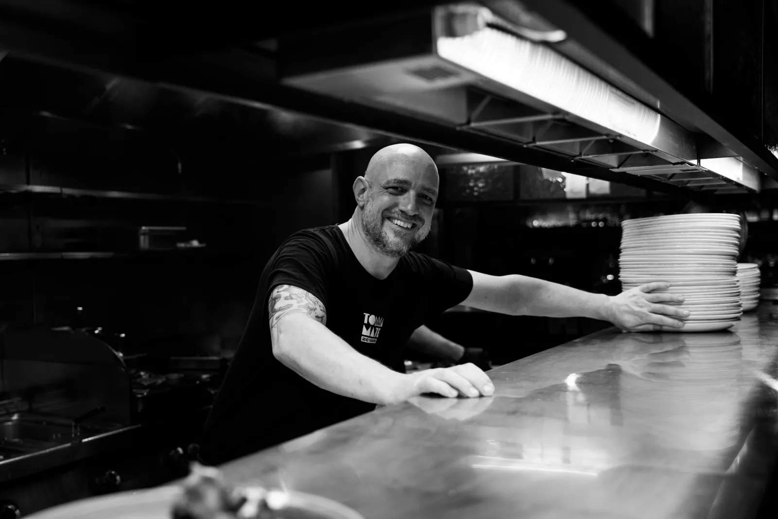 De chef leunt lachend op de keukentoog naast een stapel borden, zwart-wit